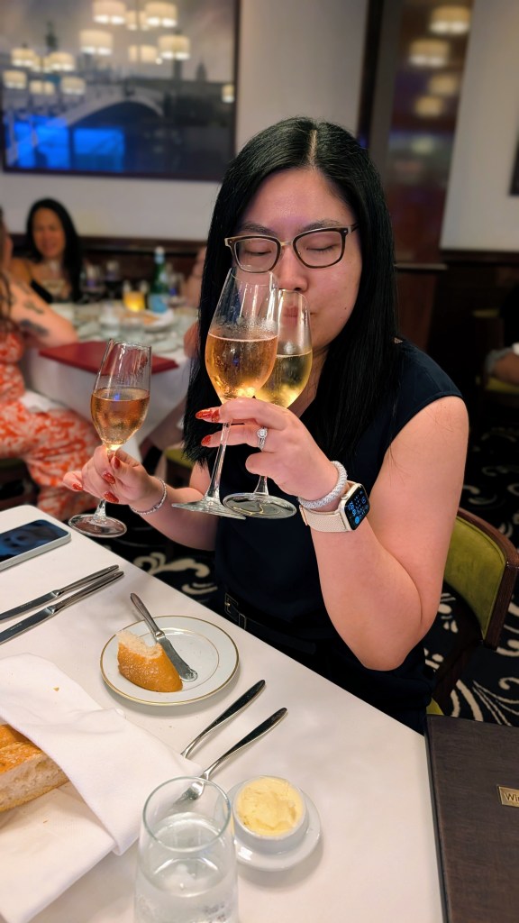 A woman wearing glasses holds two champagne glasses while smiling at the camera in a dining setting. A slice of bread and butter are on the table in front of her, along with a glass of water and utensils.